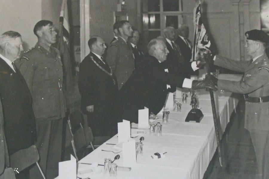 Man in military uniform passing flag to man standing behind table covered with white cloth