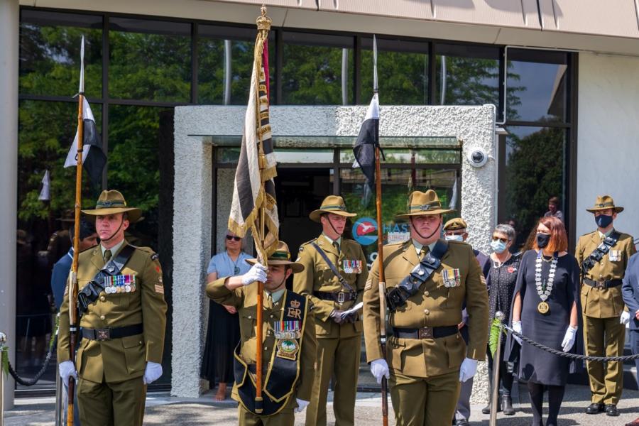 People in military uniforms carrying various flags outside building watch by people wearing masks