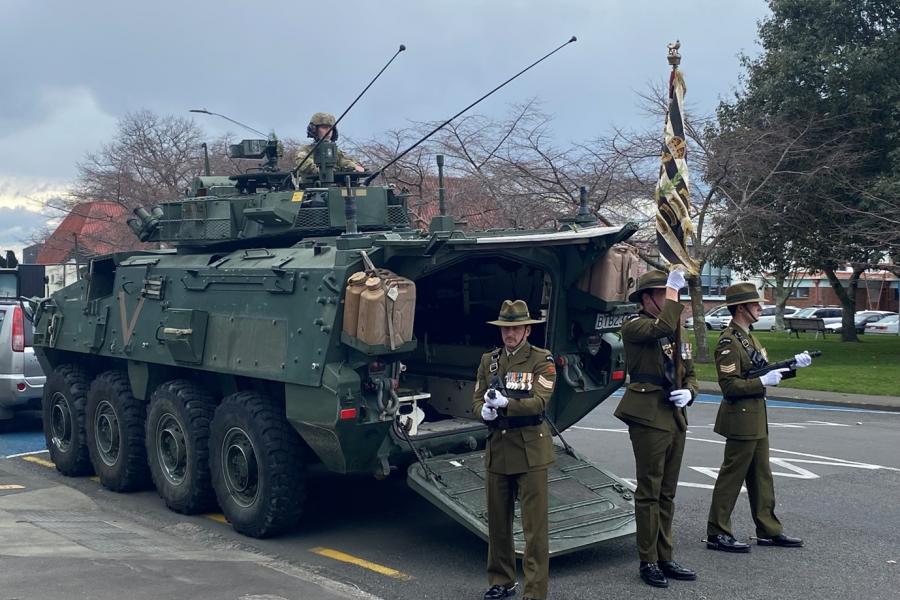 Men in military uniforms carrying flag stand beside light armoured military vehicle