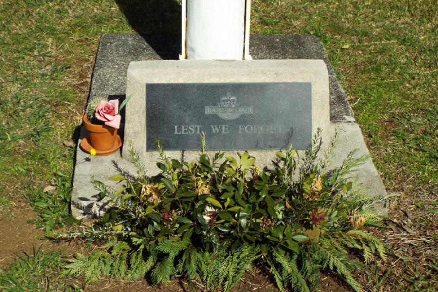 Concrete base of flagpole with marble plaque with text Lest We Forget next to floral wreath