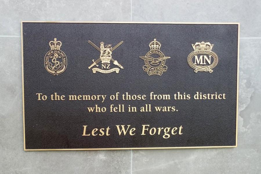 Bronze plaque with emblems of New Zealand military branches in gold under picture of a laurel wreath and heading Lest We Forget
