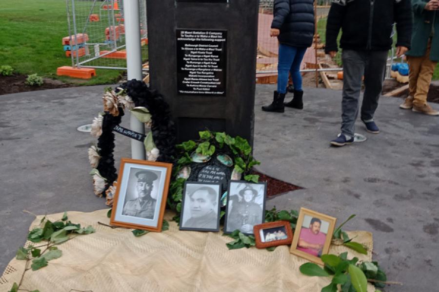 View of carved powhenua memorial to D Company 28th Māori Battalion with framed photographs of descendents laid against the bottom of the pouwhenua.