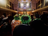 Picture of a group of people walking into the Auckland Town Hall. They are wearing 'ei katu (Cook Island wreath) made of leaves on thier head. To each side of them are members of the media holding cameras.