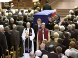 Coffin covered with New Zealand flag carried by men in uniform watched by people standing in church pews