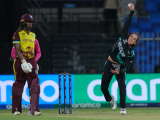 Person in black and teal sports uniform bowling a white ball next a set of cricket stumps and a person in a maroon and yellow sports uniform holding a wooden bat
