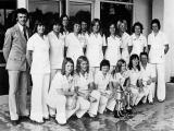 Sixteen members of a football team, all women, standing and crouching in two rows, plus one man. The women are all wearing white trouser suits. In front of them are two silvers cups decorated with ribbons.