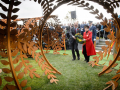 Two people approach steel sculpture holding a wreath of flowers