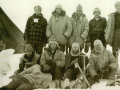 Group of ten men dressed up warmly posing for a photograph on the ice. One man lies in front while the others stand or kneel. Four ice picks are stuck in the ice in front of the group.