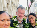 Two women each side of man wearing lei and smiling at camera