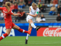 Woman in white uniform in the process of kicking a football, while a woman player in red uniform attempts to tackle her.