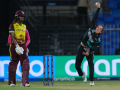 Person in black and teal sports uniform bowling a white ball next a set of cricket stumps and a person in a maroon and yellow sports uniform holding a wooden bat