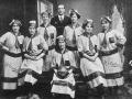 Seven women standing and seated in football uniforms, including long striped dresses and caps, one holding a football. Embroidered onto their dresses and printed on the ball is the word Polytechnic. Standing with them is a man in a suit. 