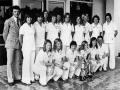 Sixteen members of a football team, all women, standing and crouching in two rows, plus one man. The women are all wearing white trouser suits. In front of them are two silvers cups decorated with ribbons.