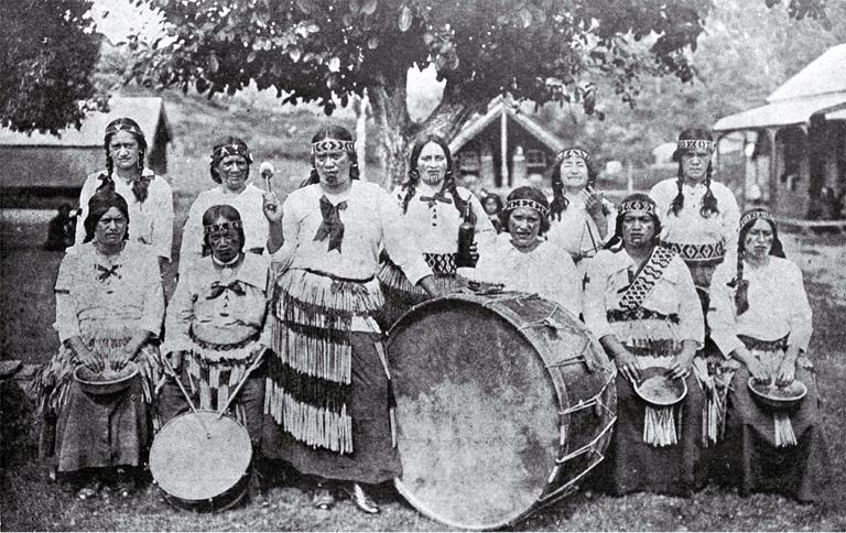 Group of Māori women wearing tīpare (headbands) and piupiu (flax skirt) in front of assorted drums
