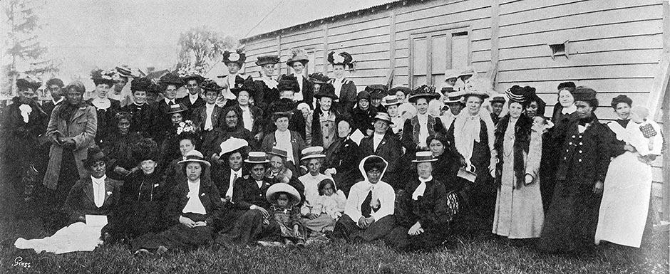 Group of women wearing various hats sitting and standing on grass outside wooden building