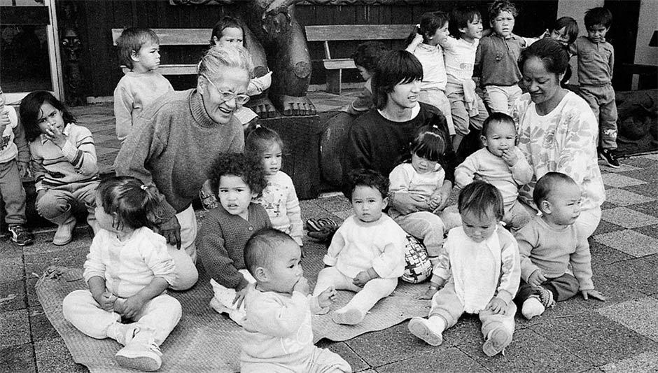 Three Maōri women holding and sitting next to a group of babies and young children