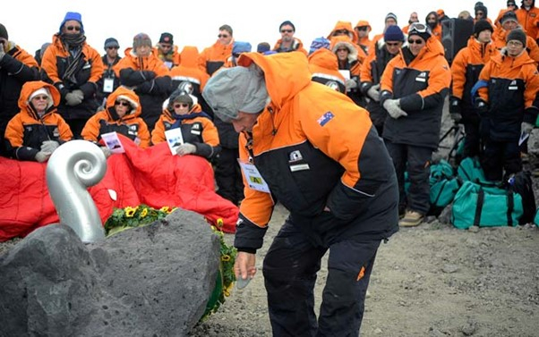 A man lays a pebble next to a memorial rock with a metal koru scupture on top. A group all dressed in thick orange and balck jackets watches on