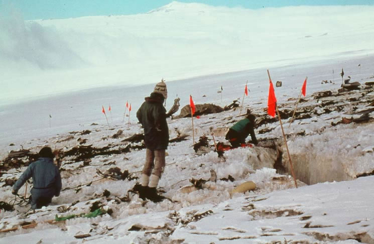 Three men on snowy slopes of mountain surround with debris. Orange flags are placed over some of the debris