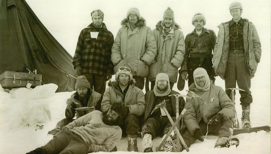 Group of ten men dressed up warmly posing for a photograph on the ice. One man lies in front while the others stand or kneel. Four ice picks are stuck in the ice in front of the group.