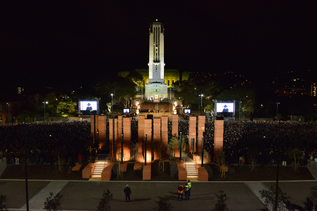 View at night down towards series of brown pillars with large crowd assembled behind them in front of tall tower