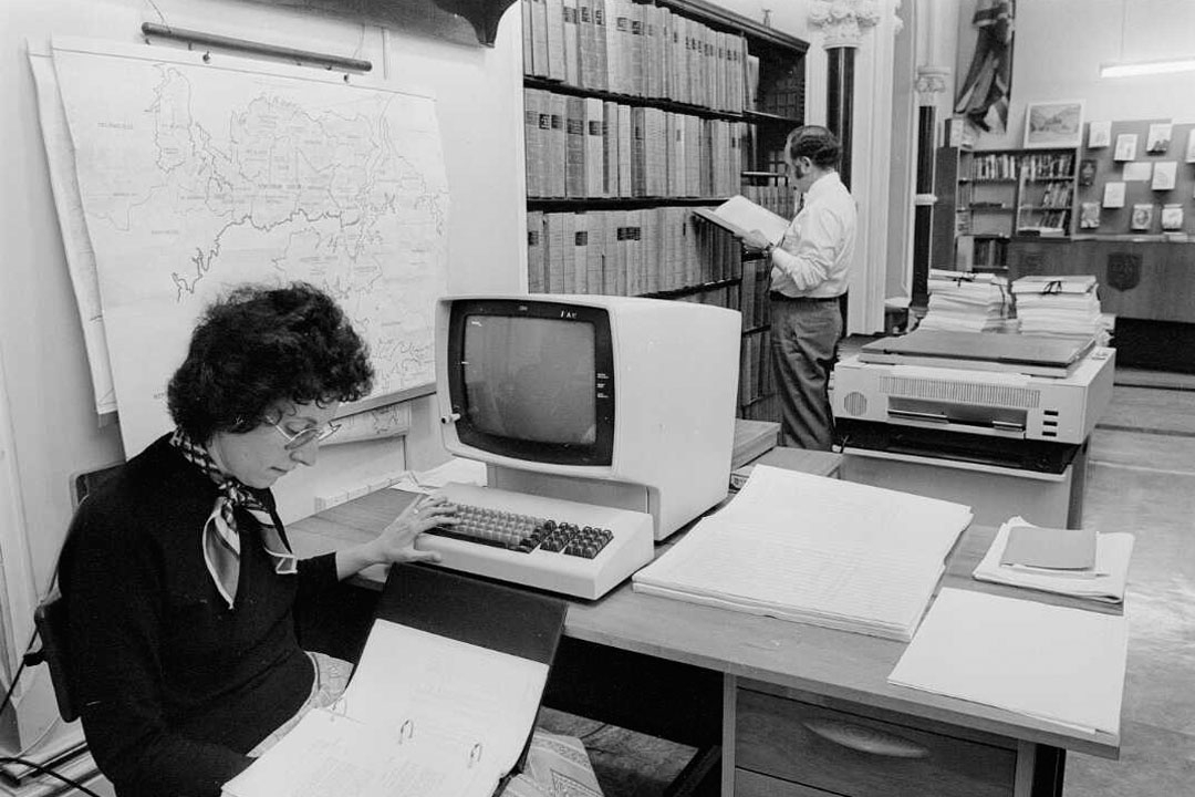 A woman sitting at a desk with a 1970s-era computer copying data from a folder 