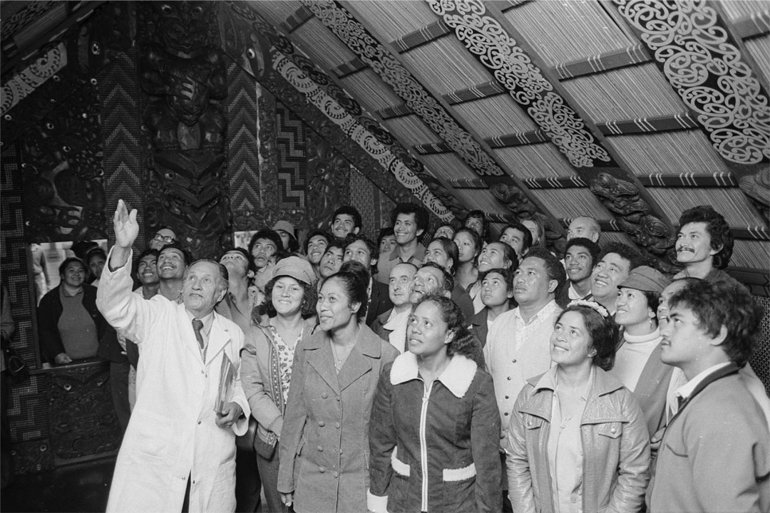 A group of people in a marae. Man in white coat in front is pointing up at ceiling.