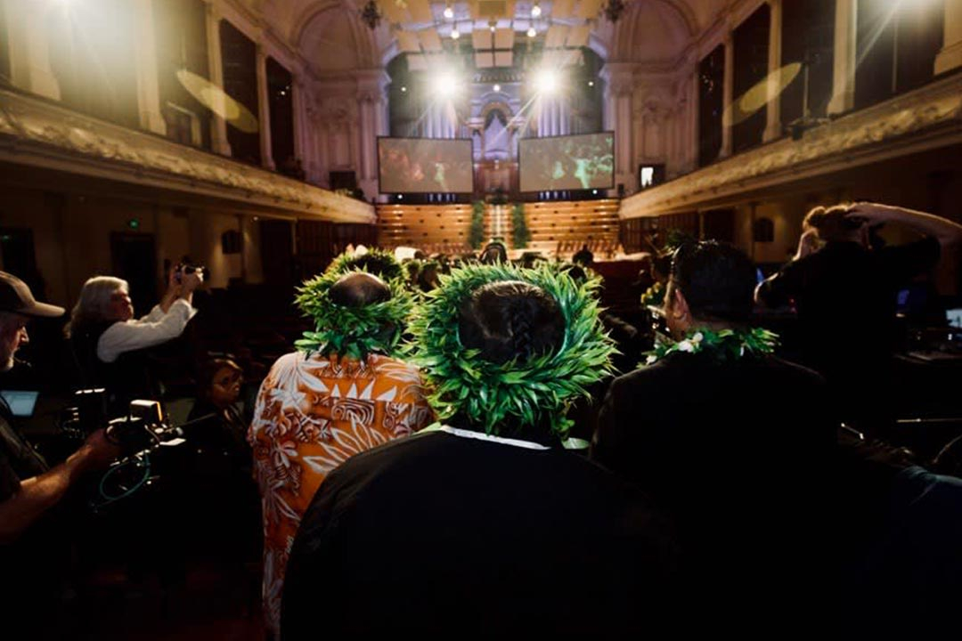 Picture of a group of people walking into the Auckland Town Hall. They are wearing 'ei katu (Cook Island wreath) made of leaves on thier head. To each side of them are members of the media holding cameras.