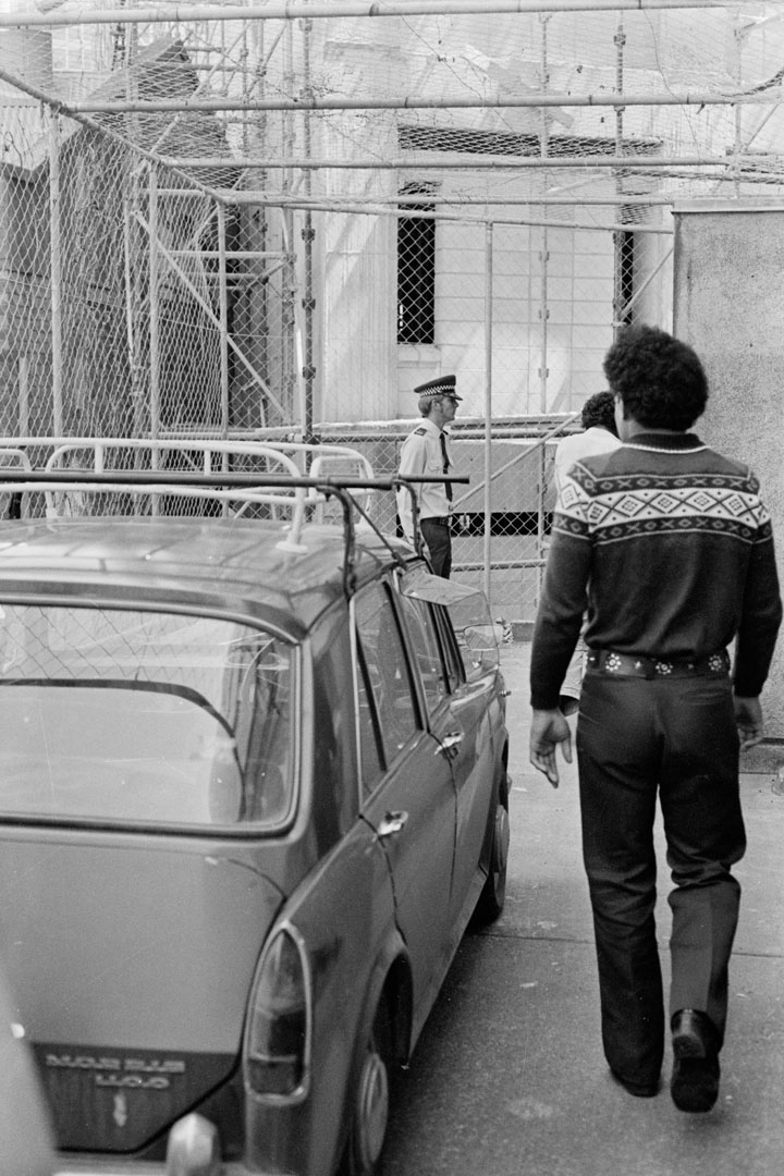 A man in a wool jumper walking next to a car, a police officer is in the background of the photo