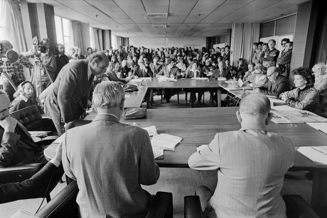 People seated around a meeting table with a large standing crowd and TV cameras watching on