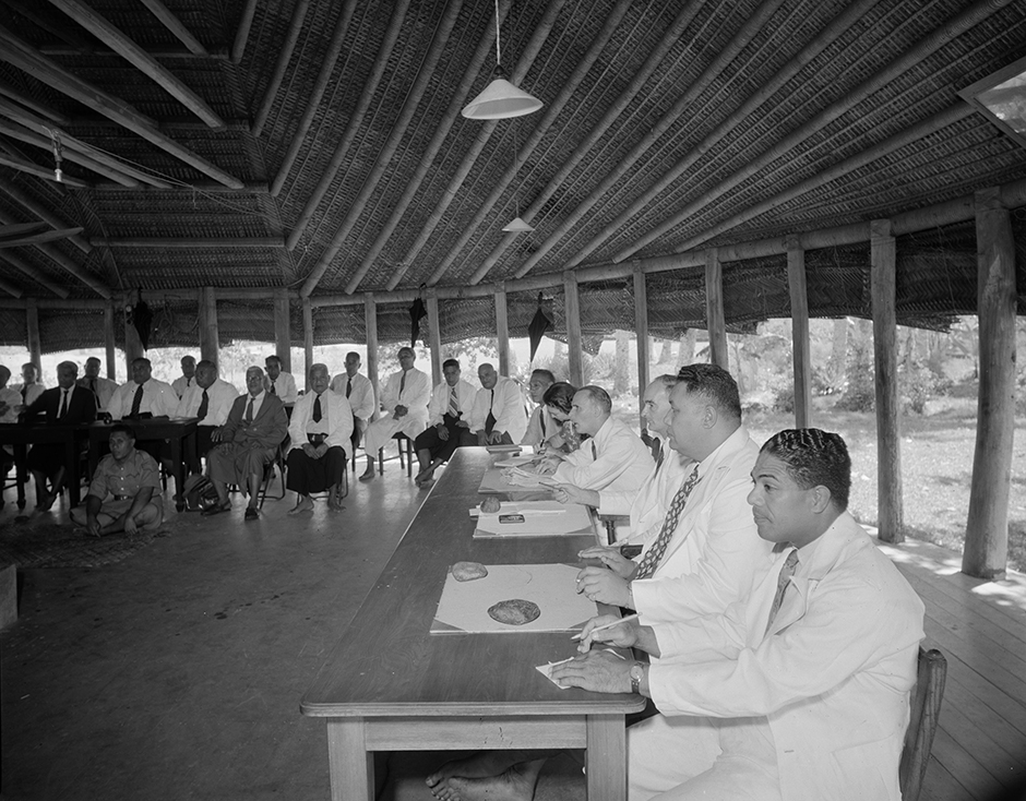 Group of men meeting in open-walled building, some share a desk while others are seated in rows.