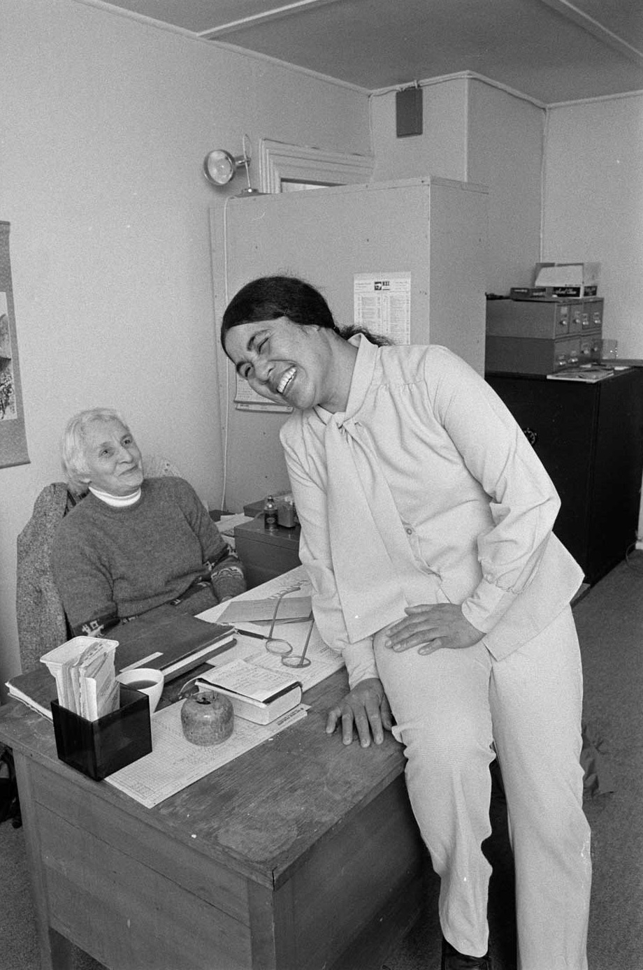 Two women laughing, one sitting behind desk and the other on top of it.