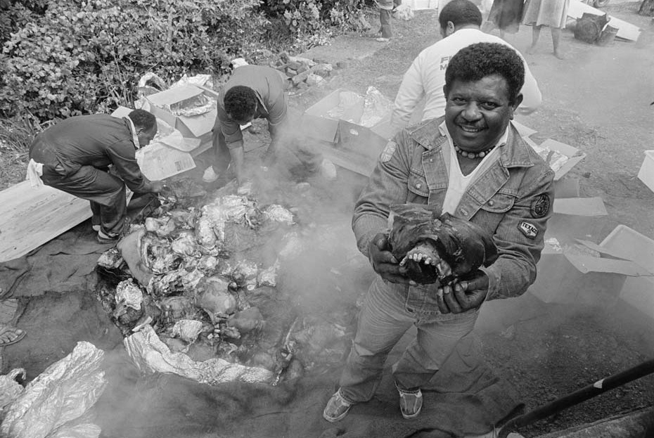 Man holding item of food taken from steaming pit filled with more food.