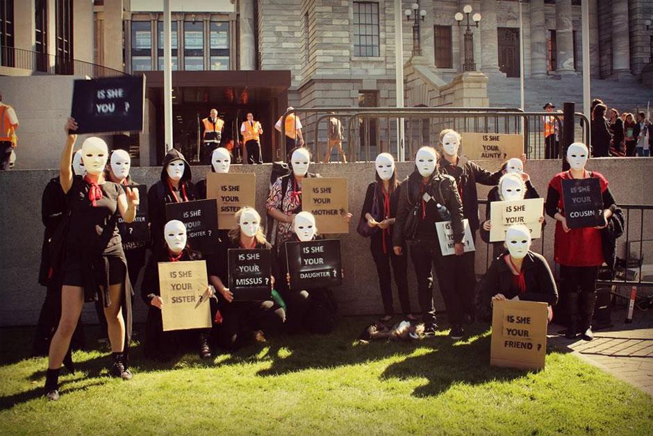 Women in white masks protesting on lawn of Parliament. One sign states, 'Is she your sister?'